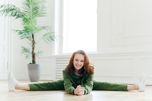 Joyful woman in green sportswear performing splits on white floor, fitness at home, bright interior with potted plant