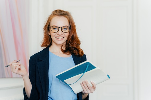 Smiling businesswoman with glasses holding a notebook and pen, professional and prepared, elegant office setting