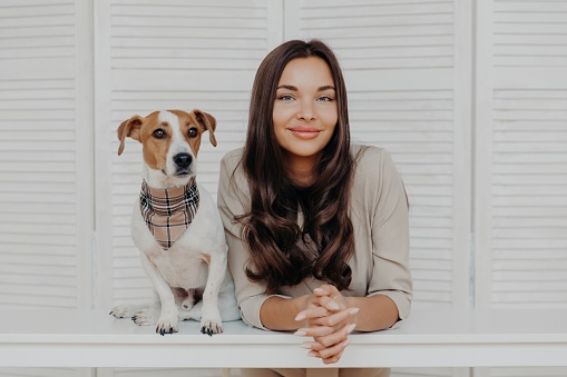 Chic woman with glossy hair beside her stylish Jack Russell Terrier against a white shuttered backdrop