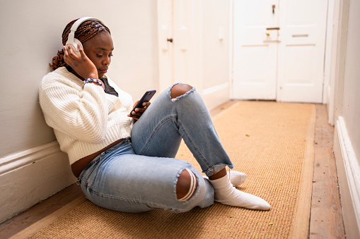 Relaxed african girl sitting on floor using mobile phone and enjoying listening to music on headphones at home