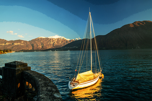 Anchored sheltering (for winter season) sailboat next to shore on lake Como.   Fantastic  amazing sunset  on Christmas holidays Snow-capped mountains in distance-italian Alps.