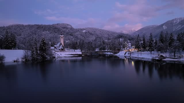 Aerial view of Bohinj lake in Slovenia at twilight in winter. Top drone view of snowy mountains, trees, church, bridge, road, city lights, houses, reflection in water, purple sky with clouds at sunset