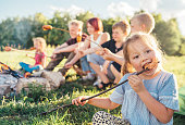 Portrait of little girl eating grilled sausage. Group of Kids - Boys and girls roasting sausages on long sticks over a campfire flame. Outdoor active time spending or camping in Nature concept.
