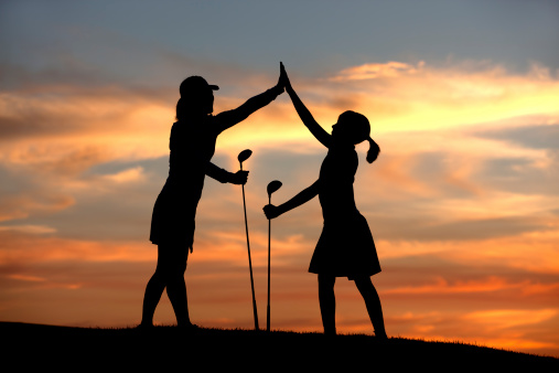 A mother and daughter in silhouette at sunset celebrate with high fives after a good drive.