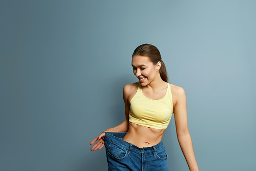 Diet concept and weight loss. Beautiful woman in oversize jeans on pastel blue background