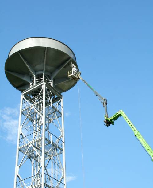 吊り上げ機のカゴに乗った作業員が、青空を背景に建設現場で金属フレームを塗装する。給水塔の修理と塗装。 - priming paint ストックフォトと画像