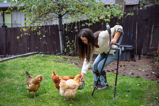 An Asian woman with a physical disability who is using crutches feeds her pet chickens in the backyard of her home.