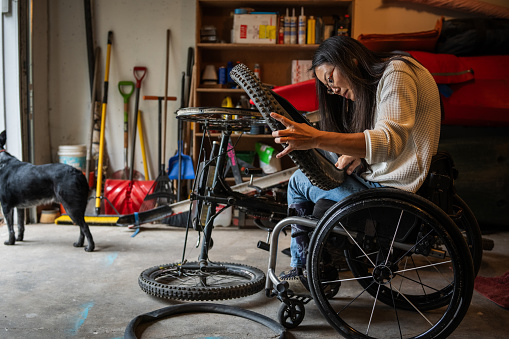 An active and adventurous young Asian woman who is a part-time wheelchair user does maintenance on the tire of her adaptive mountain bike in the garage of her home.