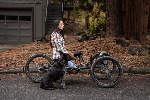 A beautiful and active Asian woman with a disability smiles and poses with her pet dog in front of her house before heading out for a ride on her adaptive mountain bike.