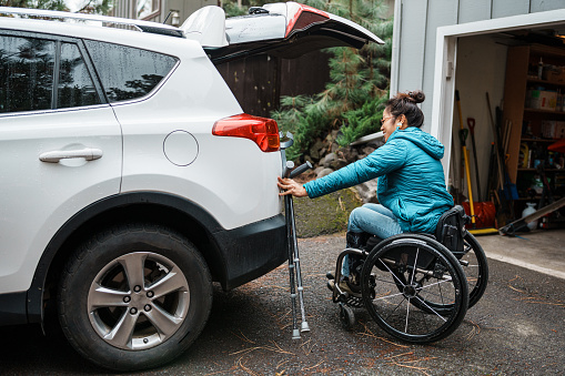 An Asian woman who is using a wheelchair places items in the open trunk of her car before driving to work and running errands.