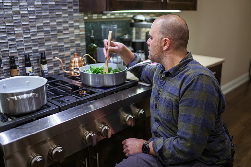 A Caucasian man who is using a wheelchair cooks dinner on the stove in the kitchen of his home.