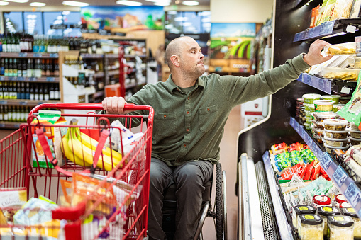 A middle aged man of Caucasian descent with a physical disability, using a wheelchair reaches to grab an item while shopping in a grocery store.