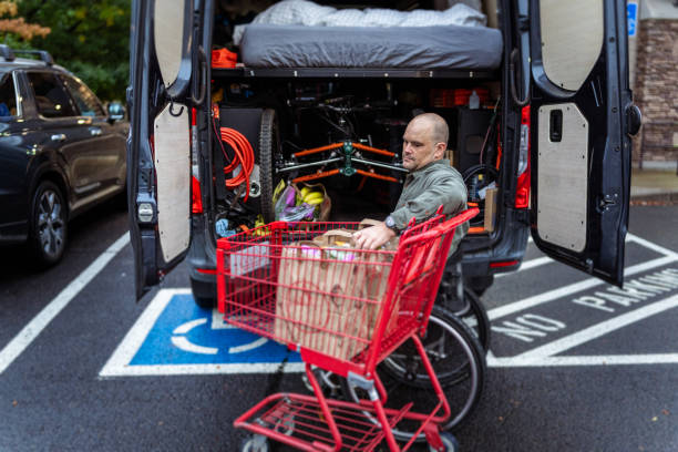 A middle aged Caucasian man using a wheelchair unloads groceries from a shopping cart, into his van in a store parking lot on an overcast day.