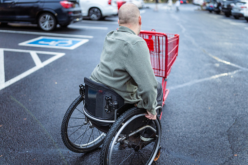 Rear view of a middle aged Caucasian man using a wheelchair pushes a red shopping cart across a parking lot on a rainy day in Central Oregon.
