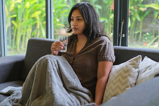 Close-up shot of sad Malaysian woman holding a glass of water while having a mental breakdown. She's sitting on a sofa in the living room with an empty stare.