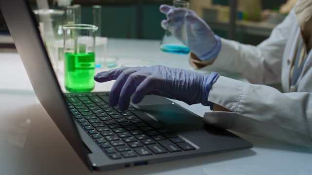 Mature Biochemist Examining Flask and Typing on Laptop in Lab