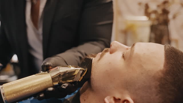 Young Barber in Suit Using Hair Clipper and Comb to Groom Customer's Beard in Salon
