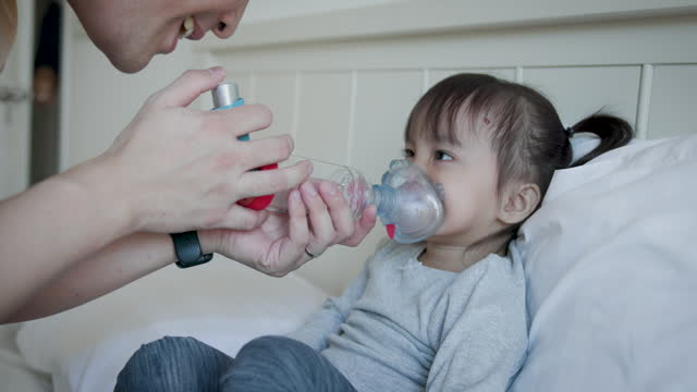 Father helps her daughter gets assistance with asthma inhaler spacer while setting in bedroom at home