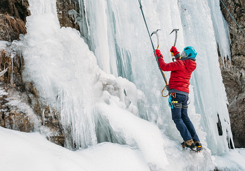 Ice climber dressed in warm winter climbing clothes, safety harness and helmet climbing frozen waterfall using two Ice climbing axes and crampons. Active people and sports activities concept image.
