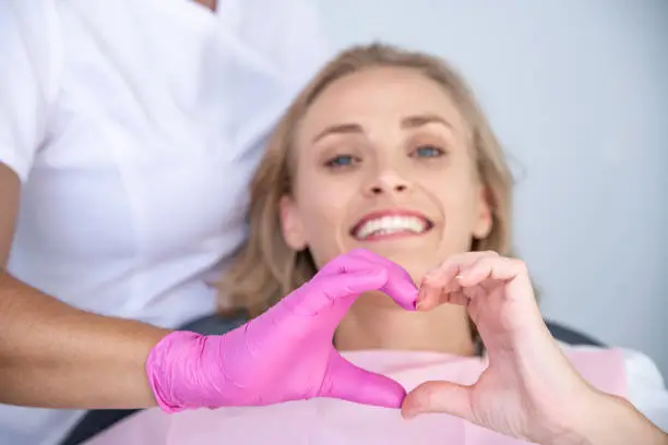 Dentist and happy smiling female patient in dental chair showing heart shape with hands. She is looking at the camera.