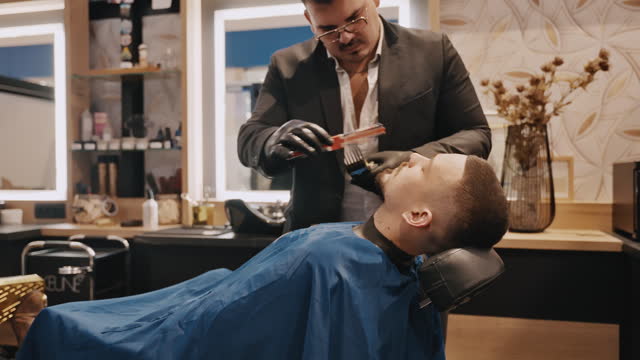 Young Barber in Suit Using Hair Clipper and Comb to trim Customer's Beard in Salon
