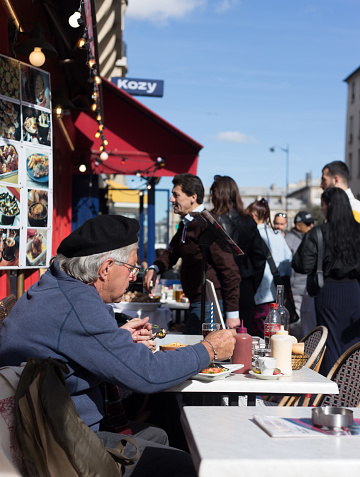 Paris, France: A senior man in a beret sits eating at a sidewalk cafe in the busy Latin Quarter.