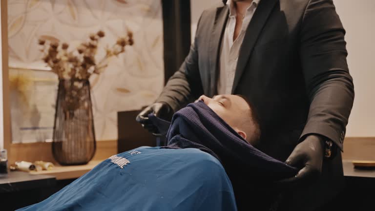 Young Barber Preparing Man For Shaving With Hot Towel on Face in Barbershop