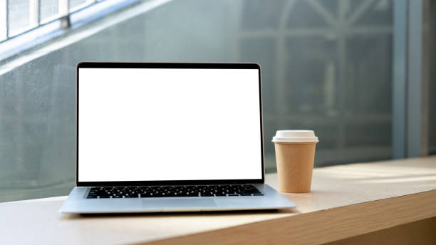 Close-up image of a white-screen laptop computer mockup and a coffee cup on a table against the window in a cafe. workplace concept