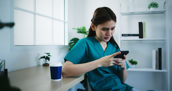Image of a young Asian nurse dressed in scrubs using a smartphone and drinking coffee in the clinic break room. Medical, people, lifestyle and relaxation concepts.