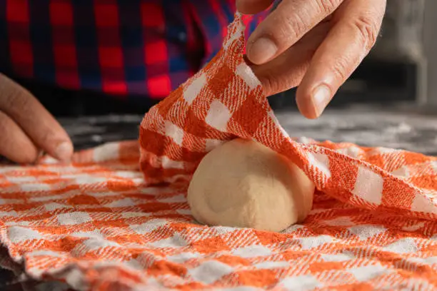 Latin man cooking a homemade, traditional and delicious pizza, chef in the kitchen with classic and vintage clothing, Wrap a ball of flour dough in a classic red square kitchen towel.