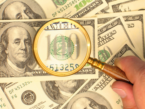 Male's hand holding a handglass and magnifying a 100-dollar note lying among others