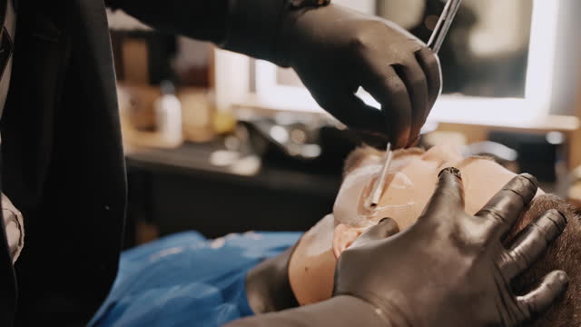 Closeup Of Hairdresser in Black Gloves Shaves Customer's Beard With Straight Razor in Salon