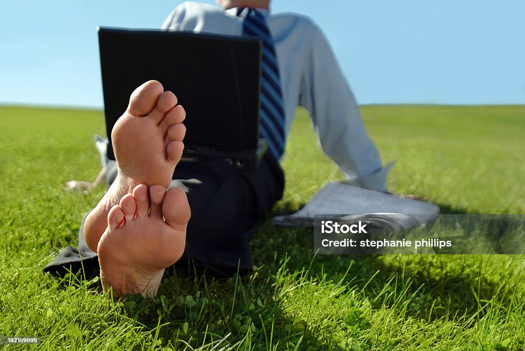 Businessman Sitting in the Grass with Bare Feet Horizontal image of a businessman taking a break on a sunny warm afternoon. He has taken his laptop outside onto a plush grass lawn. His shoes are off and he's enjoying a moment of relaxation and sun. Business Stock Photo