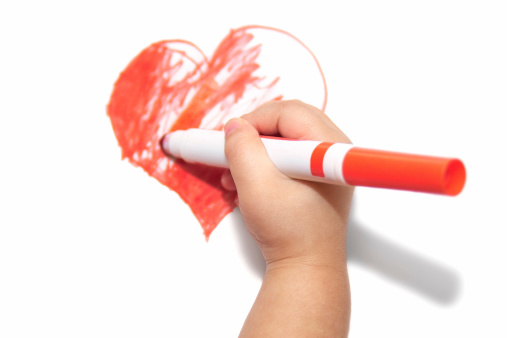 Photo of a child's hand drawing a picture of a heart with a red marker. focus is primarily on hand.