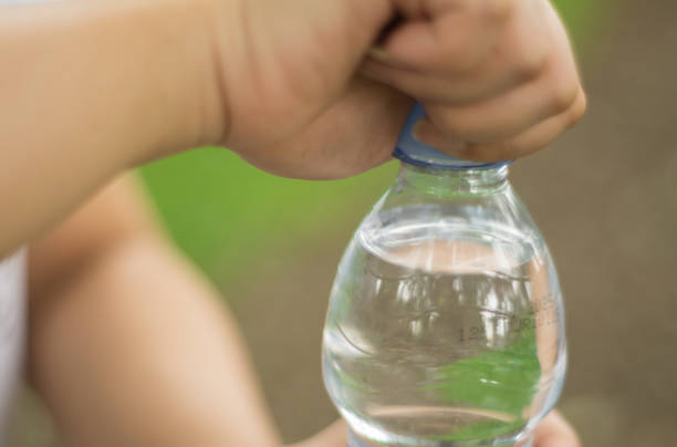 An Asian woman enjoys a refreshing sip of water from a plastic bottle while relaxing in a peaceful public park. The image captures the calm, healthy lifestyle in an outdoor setting with vibrant green trees and a well-maintained park environment. Perfect for illustrating concepts of hydration, outdoor activities, wellness, and environmental consciousness.