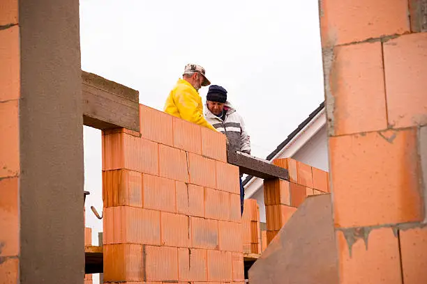 "Construction background, view through a window hole, two masons standing on a scaffolding and istalling a lintel on a structual door opening (clearspan) using a spirit level and a measuring cord for alignment. Roof and roof beam of a finished house in the background.Shot of the construction site in bad weather conditions (overcast and rain), selective focus."