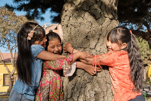three happy latina girls hugging a tree in an outdoor park in latin america bolivia