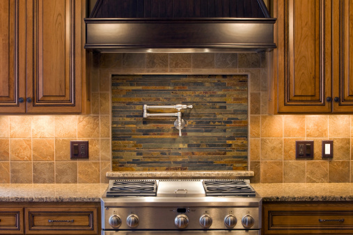 A new four burner stainless steel gas kitchen range at a high-end residential construction site is ready for its first meal. A stacked stone backsplash, pot faucet and dark stained hood are above. Cherry cabinets and granite countertops flank the left and right sides. The light reflections on the hood are from two island lights near the camera position. The two bright areas on the range are from the powerful under-hood cooking lights. http://www.banksphotos.com/LightboxBanners/Buildings.jpg