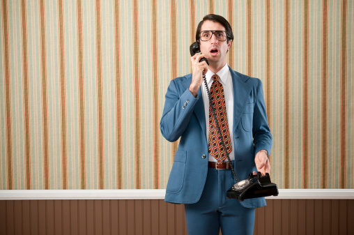 Nerdy businessman wearing a blue retro suit speaking on a black rotary vintage phone. The wall has a brown beadboard wainscoting and a striped wallpaper.Take a look at my LIGHTBOXES of other related images.
