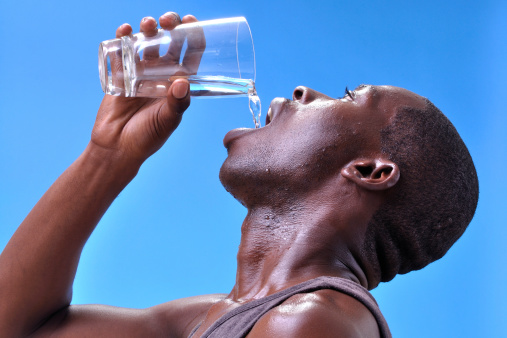 Closeup of thirsty sweaty young athletic black man pouring clean pure water into mouth from glass on blue sky background