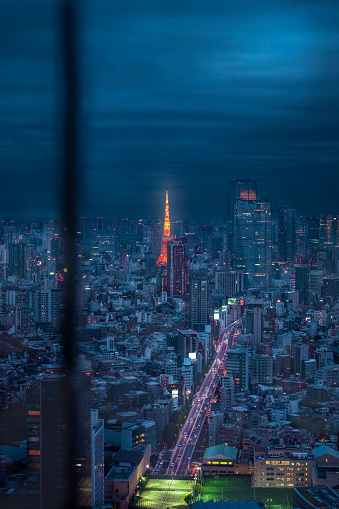 the stunning illuminated Tokyo Tower, a beacon of light amidst the nocturnal glow of Tokyo's sprawling cityscape. Shot from the observation deck of the Shibuya Sky building, the image showcases the tower radiating in its iconic orange hue, contrasting sharply with the blue and white lights of the surrounding city. Skyscrapers encroach on the skyline, their windows sparkling like stars against the dusk sky, while the streets below curve through the urban terrain, veins of light carrying the city's lifeblood. The night reveals a different kind of beauty in Tokyo, one of vibrant energy and technological wonder, with the Tokyo Tower at its heart, a symbol of Japan's post-war rebirth and modernity.
