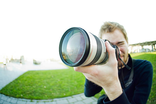 Young Professional Photographer A young adult man aims and shoots his camera armed with a telephoto zoom lens.  Fisheye /wide angle lens gives the image an interesting perspective.  Horizontal with copy space. sports photography stock pictures, royalty-free photos & images