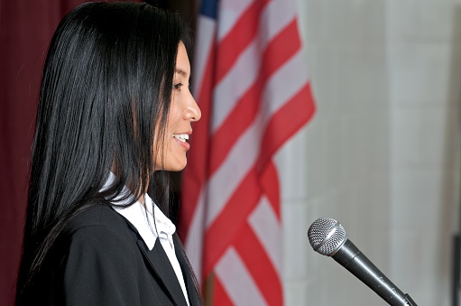 Cropped view of female politician speaking in microphone, while sitting at table near usa flag in boardroom