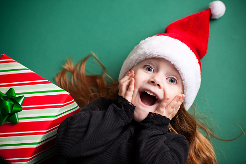 Color image of an excited 4-year-old girl wearing a Santa hat and lying on a green background.