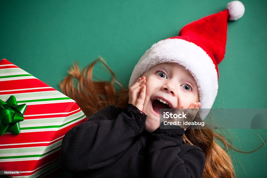 Excited Girl Wearing Santa Hat with Christmas Present Color image of an excited 4-year-old girl wearing a Santa hat and lying on a green background. Christmas Stock Photo
