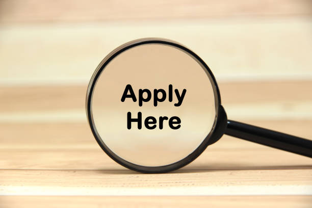 istock Apply Here text or message short word english letter on a on black magnifying glass lens brown wood grain background. 1821768467