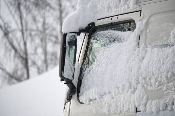 Close-up of a white truck windshield, hood, and side mirror are notably covered in thick, fluffy snow, vehicle is partially buried under the snow