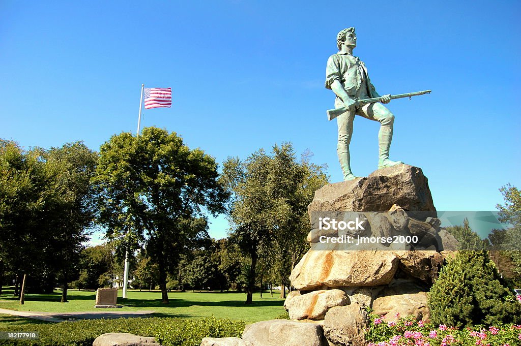 Lone statue on top of rock with American flag flying behind The famous statue of the Revolutionary War minuteman stands tall on Lexington Green. It is here the Revolutionary War started in 1775. Lexington - Massachusetts Stock Photo