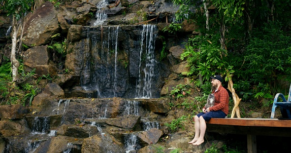 Man relaxes with cat in his arms on pier near picturesque waterfall in jungle. Relaxing in nature helps you to be alone with your thoughts. Cinematic shot of man relaxing at waterfall