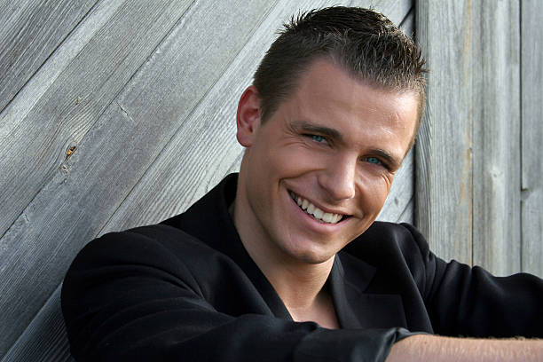 A gorgeous smiling handsome man leans against a wooden building in stark sunlight.